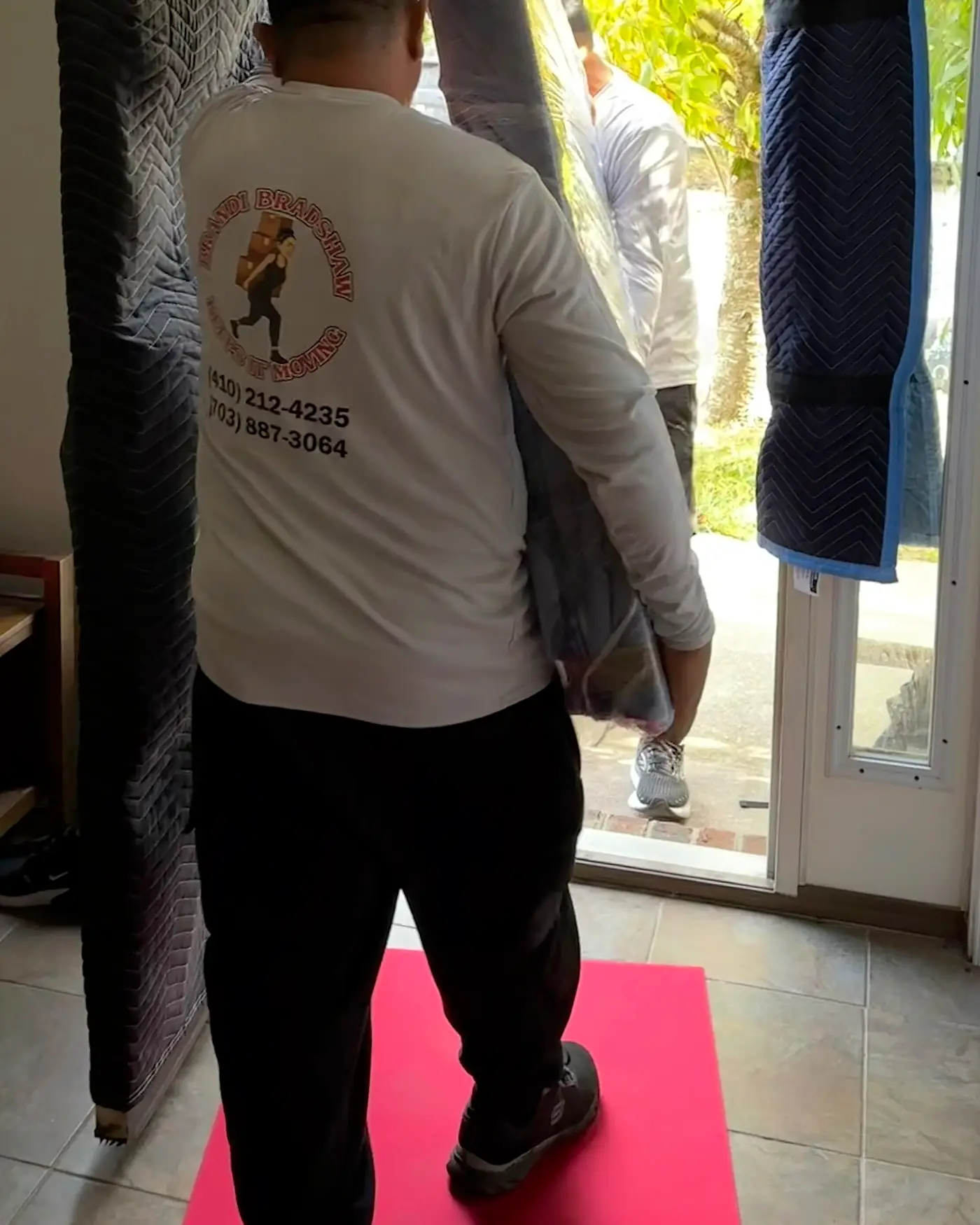 A Keeps It Moving mover carrying a wrapped mattress through a padded doorway, floor runner down to protect the home